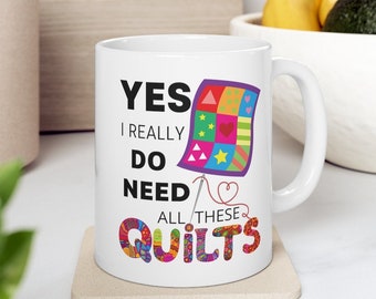 Op de afbeelding: Witte keramische mok met een zwart en kleurrijk ontwerp. De mok heeft de tekst "YES I REALLY DO NEED ALL THESE QUILTS" met een kleurrijk quiltblok en een naald en draad.