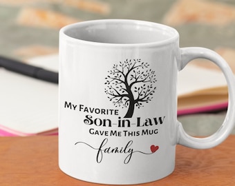 May include: White ceramic mug with a black tree design and the text "My Favorite Son-in-Law Gave Me This Mug family" in black script. A small red heart is next to the word "family". The mug has a curved handle.