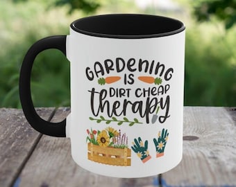 May include: White ceramic mug with a black rim and handle. The mug has a gardening themed design with the text "Gardening is dirt cheap therapy" and illustrations of carrots, flowers, and gardening gloves.