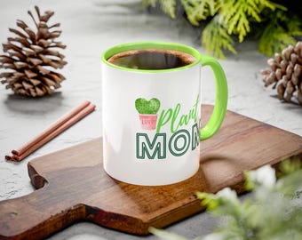 May include: White ceramic mug with a green rim and handle. The mug has a green cactus illustration and the text "Plant Mom" in black.