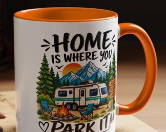 Op de afbeelding: Een witte keramische mok met een oranje binnenkant en handvat, voorzien van een kleurrijke afbeelding van een camper, kampvuur, dennenbomen en bergen. De tekst luidt "HOME IS WHERE YOU PARK IT", met kleine hartjes en een wegwijzer.