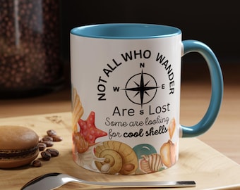 May include: A white ceramic mug with a blue interior and handle, decorated with seashells and the text "NOT ALL WHO WANDER ARE LOST." A compass rose graphic is also featured. A spoon and a macaron are in the foreground.