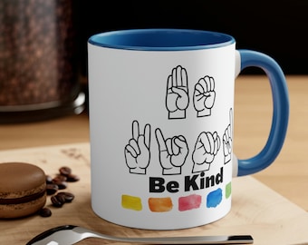 May include: White ceramic mug with a blue rim and handle. The mug features a design of hands signing "Be Kind" in American Sign Language. The text "Be Kind" is printed below the hands in black. The background of the mug is white with a watercolor rainbow design at the bottom.