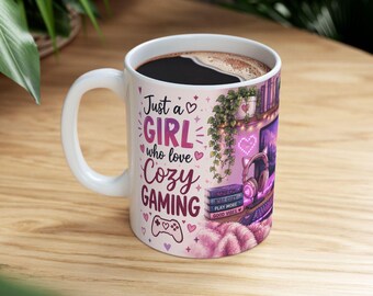Op de afbeelding: Een witte keramische mok met de tekst Just a GIRL who love COZY GAMING in roze en paarse letters. Het ontwerp toont een gaming-opstelling met een monitor, koptelefoon met kattenoren, een stapel boeken, een hangplant en een gamecontroller-icoon.