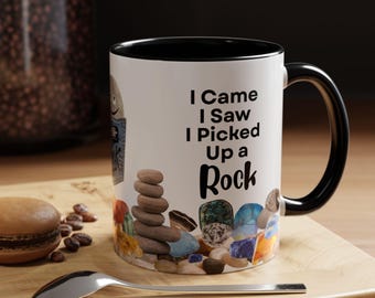 May include: White ceramic mug with a black interior and handle. The mug features the text "I Came I Saw I Picked Up a Rock" in black. The mug is decorated with a variety of colorful rocks and stones.
