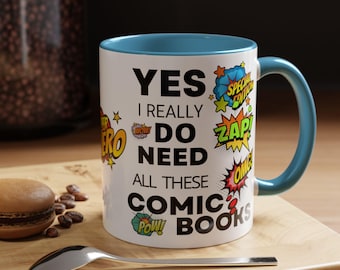 May include: A white ceramic mug with a blue interior and handle, featuring comic book-style text that reads "YES I REALLY DO NEED ALL THESE COMIC BOOKS." The mug is decorated with comic book sound effects and starbursts.