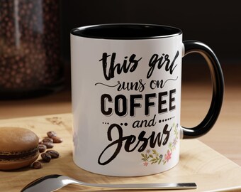 Op de afbeelding: Een witte keramische mok met een zwarte binnenkant en handvat, voorzien van de zwarte tekst "this girl runs on COFFEE and Jesus" in een mix van script- en blokletters, geaccentueerd door een kleine kleurrijke bloemengrafiek rechtsonder.
