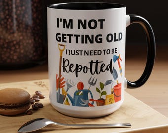 May include: White ceramic mug with a black interior and handle. The mug features the text "I'M NOT GETTING OLD I JUST NEED TO BE Repotted" with gardening tools illustrations. A spoon and a pastry are on a wooden surface.