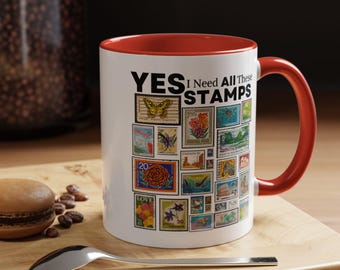 Op de afbeelding: Witte keramische mok met een rode binnenkant en handvat, versierd met een collage van kleurrijke postzegels. De mok heeft de tekst "YES I Need All These STAMPS" in het zwart. Een lepel en een macaron liggen op een houten oppervlak.