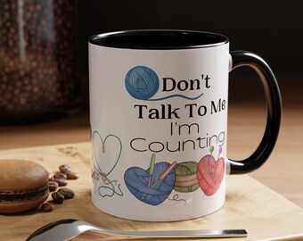 May include: White ceramic mug with a black rim and handle. The mug has a design of yarn balls, needles, and the text "Don't Talk To Me I'm Counting".