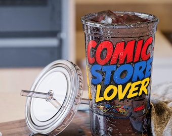 Op de afbeelding: Een doorzichtige plastic beker gevuld met een donkere vloeistof en ijs, voorzien van de tekst COMIC STORE LOVER in dikke, rood-blauw-gele stripboekletters. De beker bevat een bijpassend doorzichtig deksel met een rietje en een stripboekafbeelding.