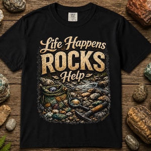 以下が含まれることがあります： Life Happens ROCKS Helpというテキストがプリントされた黒の半袖Tシャツ。デザインには、色とりどりの石、編み込みのバッグ、地質調査用ハンマーが描かれています。Tシャツはクルーネックで、標準的なフィット感です。
