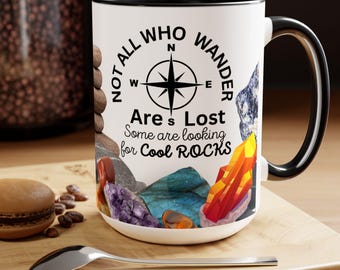 May include: White ceramic mug with a black rim and handle. The mug features a black and white compass design with the text "Not all who wander are lost. Some are looking for cool rocks." and a variety of colorful rocks.