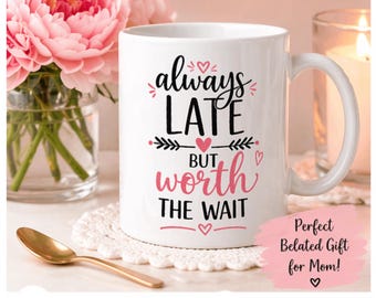 May include: White ceramic mug with the text "always late but worth the wait" in black and pink script. The mug is decorated with hearts and floral accents. A gold spoon and pink flowers are in the background.