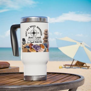 May include: White travel mug with a black lid and handle. The mug has a compass and rocks printed on it. The text on the mug reads "Not all who wander are lost. Some are looking for cool rocks."