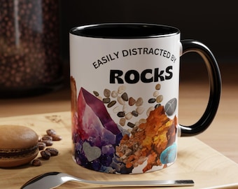 May include: White ceramic mug with a black rim and handle. The mug has a graphic of colorful rocks and the text "Easily Distracted by Rocks".