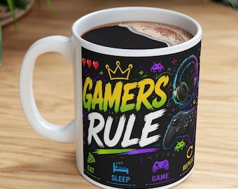 Op de afbeelding: Een witte keramische mok met een zwarte achtergrond en de kleurrijke tekst GAMERS RULE. Het ontwerp bevat een gele kroon, rode harten, een gaming-headset, een controller en iconen voor eten, slapen, gamen en herhalen in felle neonkleuren.