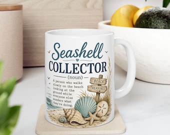 Op de afbeelding: Een witte keramische mok met de tekst Seashell Collector (noun) en een definitie over wandelen op het strand. Het ontwerp bevat illustraties van diverse schelpen, een zeester, een zanddollar en een houten bordje met de tekst Beach Treasures.