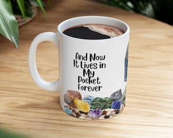 Op de afbeelding: Witte keramische koffiemok met de tekst "And Now It Lives in My Pocket Forever." De mok is gevuld met donkere koffie en versierd met kleurrijke edelstenen. De mok staat op een houten oppervlak.
