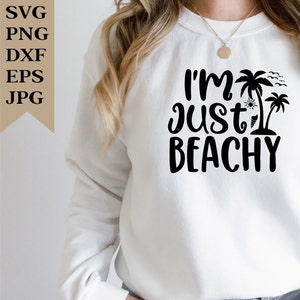 May include: A white sweatshirt with the text "I'm Just Beachy" in black lettering. The text is surrounded by palm trees, a sun, and a beach umbrella.
