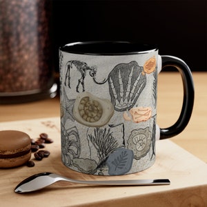 May include: A black and white ceramic coffee mug with a black handle. The mug features a pattern of various fossils, including a trilobite, ammonite, and a dinosaur bone.