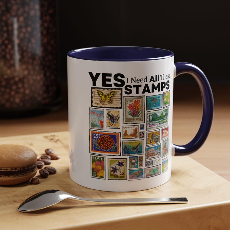May include: White ceramic mug with a navy blue interior and handle, decorated with a collage of colorful postage stamps. The mug has the text "YES I Need All These STAMPS" in black letters.