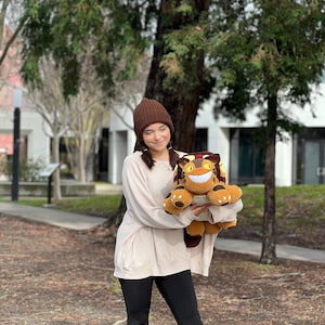 May include: A person wearing a brown beanie and a white sweater is holding a crocheted plush toy cat. The cat is brown and black with yellow eyes and a wide smile. The person is standing in front of a green tree.