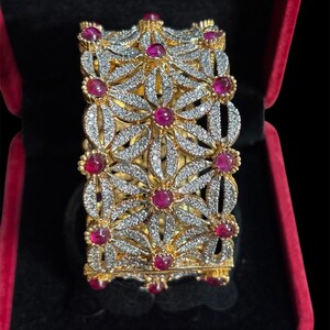 May include: A gold-toned bracelet with a floral design, set with clear crystals and vibrant red gemstones. The bracelet is rectangular and sits inside a red velvet jewelry box, against a black background.