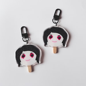 May include: Two clear acrylic keychains shaped like popsicle faces with black hair, red eyes, and pink smiles. Each keychain has a wooden stick and a black metal clasp. The faces have a glittery, sparkly appearance.