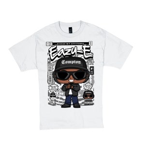 Puede incluir: Camiseta blanca con un estampado gráfico en blanco y negro de un rapero de dibujos animados con gafas de sol y una gorra de béisbol negra. El gráfico incluye el texto "Eazy-E", "Compton", "Ruthless Family" y "#1 First Issue".