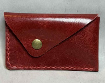 Op de afbeelding: Een kleine, rechthoekige, rode leren kaarthouder met een klepsluiting. De klep is vastgezet met een goudkleurige knoop. De randen zijn gestikt met rode draad. De kaarthouder is ontworpen om visitekaartjes of creditcards in op te bergen.