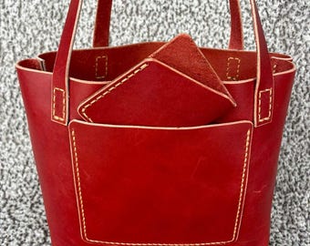 Op de afbeelding: Een rode leren tote bag met een bijpassende portemonnee. De tas heeft een rechthoekige vorm met een voorvak en twee handvatten. De portemonnee is driehoekig en past in de tas. De stiksels zijn in een contrasterende crèmekleur.