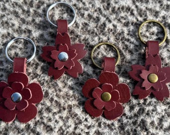 Op de afbeelding: Vier bordeauxrode leren bloemvormige sleutelhangers met metalen ringen. Elke bloem heeft een ander metalen midden, waaronder zilver- en goudkleurig. De sleutelhangers zijn gerangschikt op een getextureerd, neutraal gekleurd oppervlak.