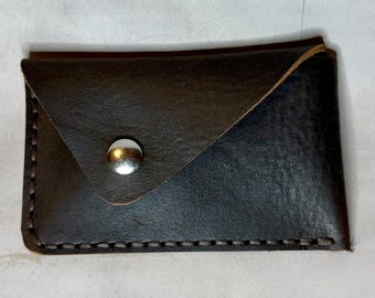 Op de afbeelding: Een donkerbruine leren kaarthouder met een zilveren drukknoopsluiting. De randen zijn gestikt met wit garen. De kaarthouder is rechthoekig met een driehoekige flap.