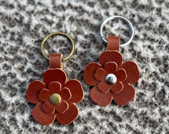 Op de afbeelding: Twee bruine leren bloemvormige sleutelhangers. Eén heeft een messing midden en de andere een zilveren midden. Elke sleutelhanger heeft een kleine leren lus bevestigd aan een metalen ring.