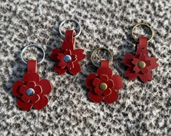 Op de afbeelding: Vier rode leren bloemvormige sleutelhangers met zilver- en messingkleurige hardware. Elke sleutelhanger heeft een bloemontwerp met een centrale knop. De sleutelhangers zijn gerangschikt op een getextureerd grijs oppervlak.