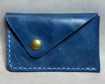 Op de afbeelding: Een kleine, blauwe leren kaarthouder met een klepsluiting en een messing drukknop. De randen zijn gestikt met lichtblauw garen. De portemonnee is rechthoekig en lijkt handgemaakt.