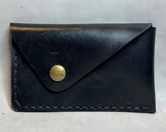 Op de afbeelding: Een zwarte leren kaarthouder met een klepsluiting en een goudkleurige knop. De randen zijn gestikt met een contrasterend draad. De portemonnee is rechthoekig en lijkt handgemaakt.