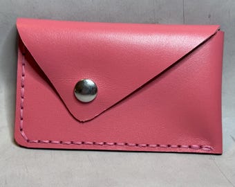 Op de afbeelding: Een roze leren kaarthouder met een flap en een zilveren drukknop. De randen zijn gestikt met roze draad. De rechthoekige kaarthouder lijkt handgemaakt.