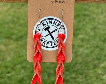 Op de afbeelding: Rode gevlochten leren oorbellen hangen aan een bruin label. Op het label staat "Handmade with love" en het heeft een cirkelvormig logo met de tekst "KINNEY CRAFTED" en een hamer en gekruiste gereedschappen. De oorbellen hangen aan zilveren haken.