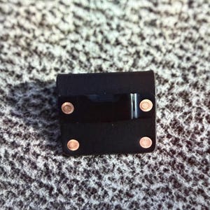 May include: A small, square black leather case with four copper-colored rivets and a shiny black band across the front. The case appears to be closed and is sitting on a textured, gray surface.