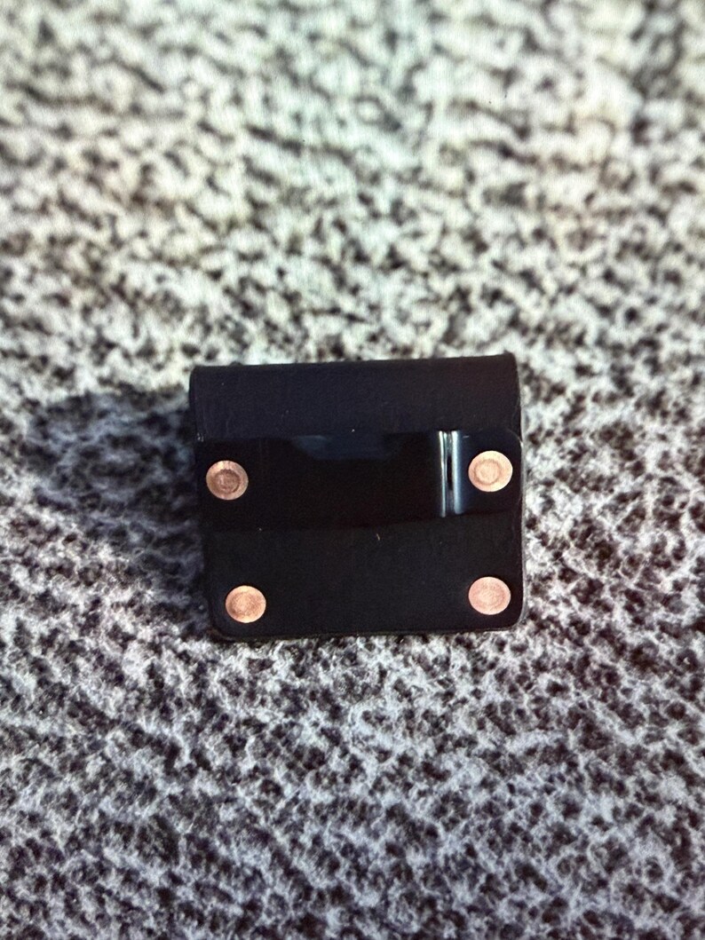 May include: A small, square black leather case with four copper-colored rivets and a shiny black band across the front. The case appears to be closed and is sitting on a textured, gray surface.