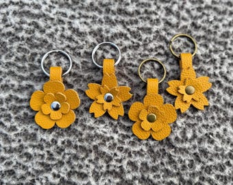 Op de afbeelding: Vier gele leren bloemvormige sleutelhangers met zilveren en messing sleutelringen. Elke bloem heeft een ander middendetail, inclusief zilveren en messing klinknagels. De sleutelhangers zijn gerangschikt op een getextureerd, grijs oppervlak.