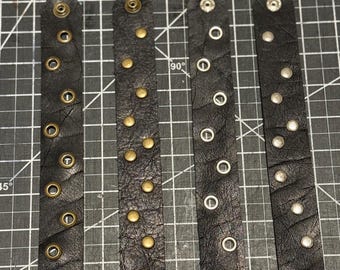Op de afbeelding: Vier zwarte leren polsbandjes met metalen studs en oogjes. De polsbandjes hebben verschillende stijlen metalen hardware, waaronder messing en zilverkleurige klinknagels en oogjes. Het leer lijkt getextureerd.