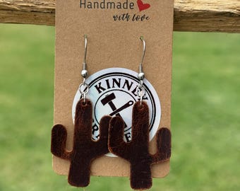 Op de afbeelding: Bruin leren cactus oorbellen die aan zilveren haken hangen. De oorbellen worden gepresenteerd op een bruin label met de tekst "Handmade with love". Een witte sticker met de tekst "KINNIE" is zichtbaar.