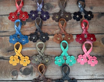 Op de afbeelding: Een verzameling leren bloemvormige accessoires in verschillende kleuren, waaronder rood, blauw, geel en roze. Elk accessoire heeft een lusvormige band en is versierd met bloemvormige versieringen. De accessoires worden gepresenteerd op een houten oppervlak.