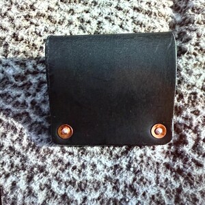 May include: A black leather wallet with copper rivets. The wallet is rectangular with a curved top and is closed. The rivets are on the bottom corners of the wallet.