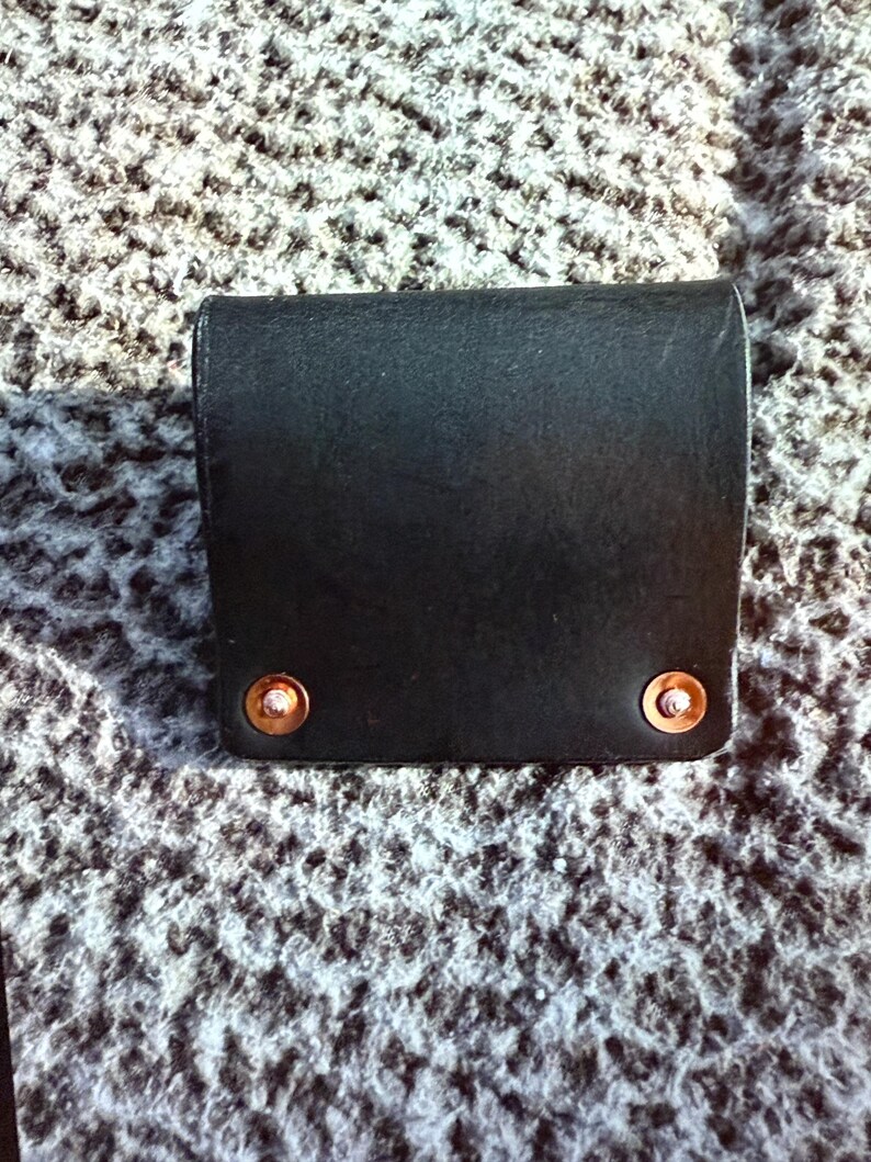 May include: A black leather wallet with copper rivets. The wallet is rectangular with a curved top and is closed. The rivets are on the bottom corners of the wallet.