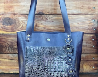 Op de afbeelding: Een donkerpaarse leren tote bag met een voorvak met krokodillenleerpatroon. De tas heeft lange hengsels, gouden klinknagels en een bloemenbedel. De tas wordt getoond tegen een houten achtergrond.