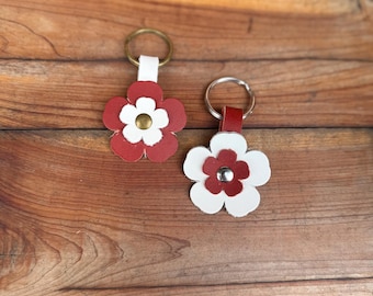 Op de afbeelding: Twee bloemvormige sleutelhangers. De ene sleutelhanger heeft een rode bloem met witte bloemblaadjes en een goudkleurig centrum. De andere heeft een witte bloem met rode bloemblaadjes en een zilverkleurig centrum. Beide hebben een leren bandje en een metalen ring.